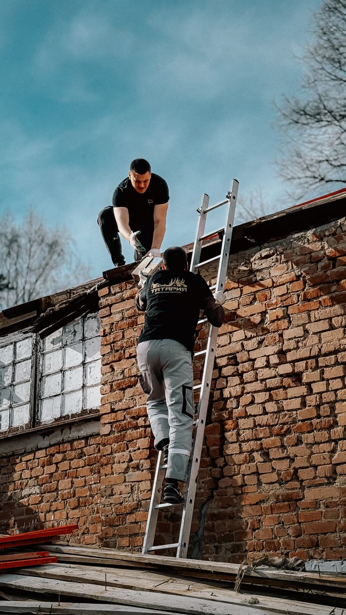 services-01 Two construction workers using a ladder to reach a brick building roof under a blue sky.