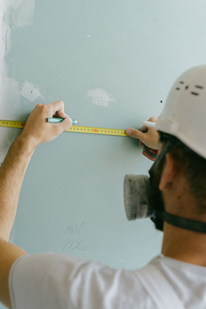 A builder in a hard hat measures a wall's dimensions with a tape measure.