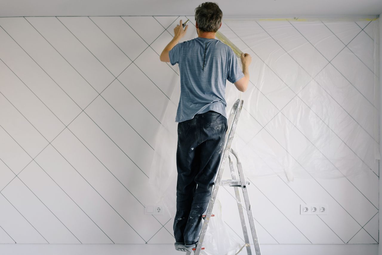 Full body back view of faceless male in shirt and dirty pants repainting wall while standing on ladder in bright empty room in daytime