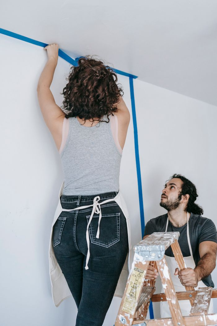 contact-img A couple working on a home renovation project using painter's tape and a ladder indoors.